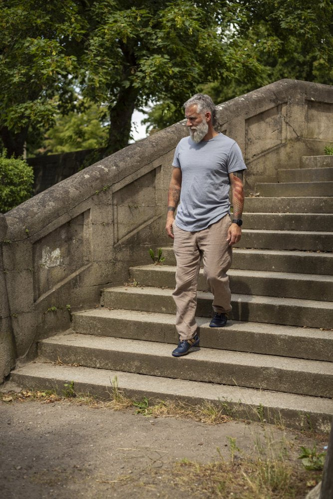 Mężczyzna z siwą brodą i tatuażami schodzi po betonowych schodach w parku. Nosi jasnoszarą koszulkę, beżowe spodnie i buty ZAQQ QANBERRA Navy barefoot z minimalną podeszwą. W tle widać zielone drzewa i krzewy.