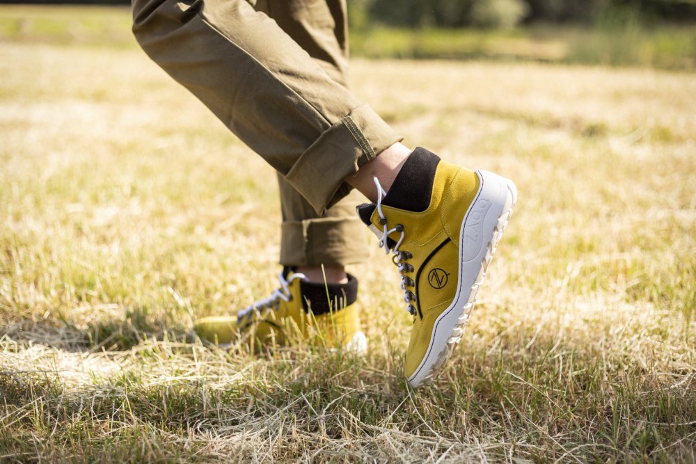 Osoba nosząca TERRAQ Yellow Waterproof Sneakers od ZAQQ, z wysokimi cholewkami, białymi podeszwami i ciemnymi sznurówkami, stoi na suchej trawie. Ma podwinięte spodnie khaki przed słonecznym tłem na zewnątrz, które doskonale eksponuje te solidne buty outdoorowe.