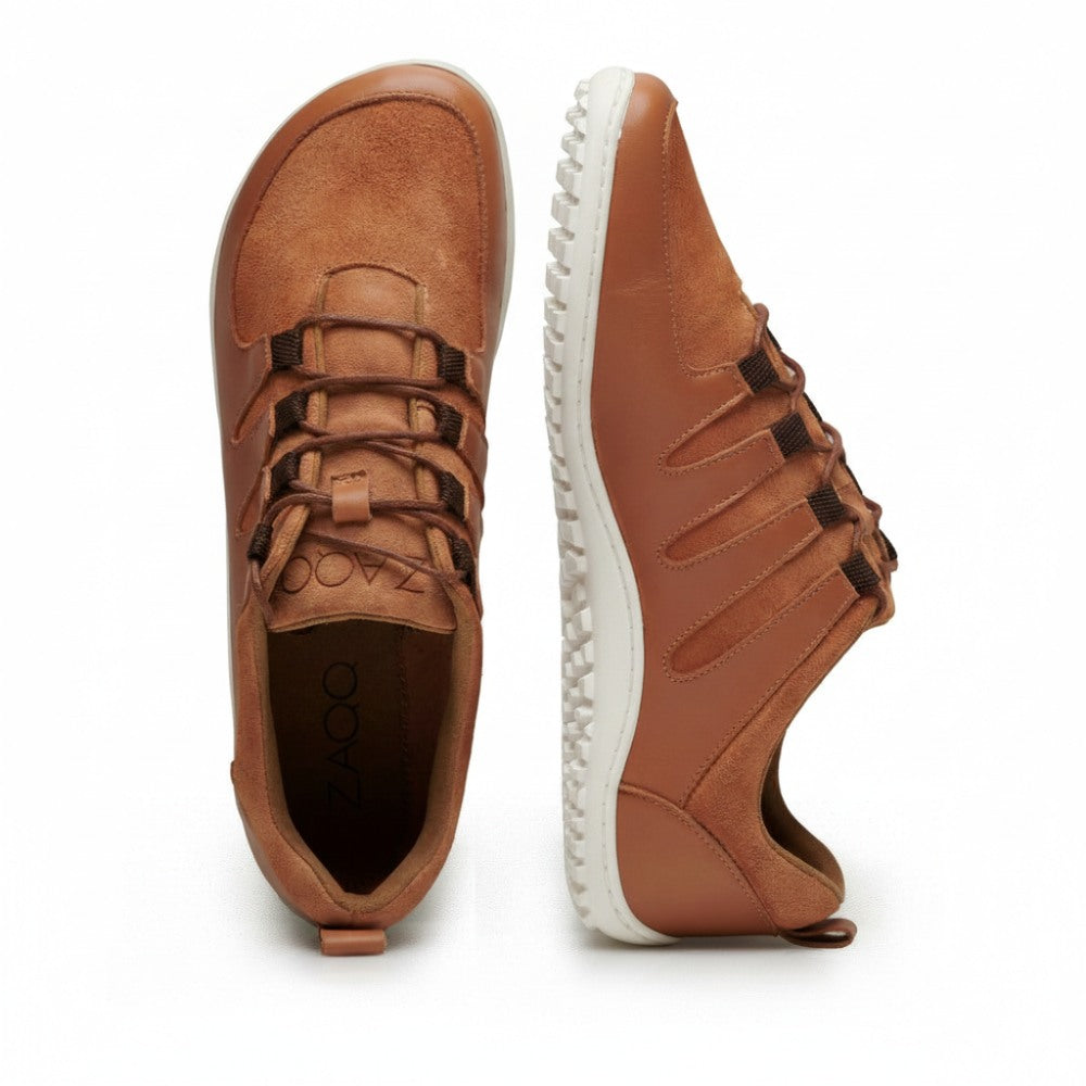 Zwei SHOQQ-Cognac-Sneaker von ZAQQ werden vorgestellt. Einer ist aufrecht positioniert, um die Draufsicht zu zeigen, während der andere auf der Seite liegt und das Design der strukturierten weißen Sohle zeigt. Diese Barfußschuhe haben Schnürfronten und ein sichtbares Branding im Inneren eines Schuhs, was ihren außergewöhnlichen Tragekomfort unterstreicht.